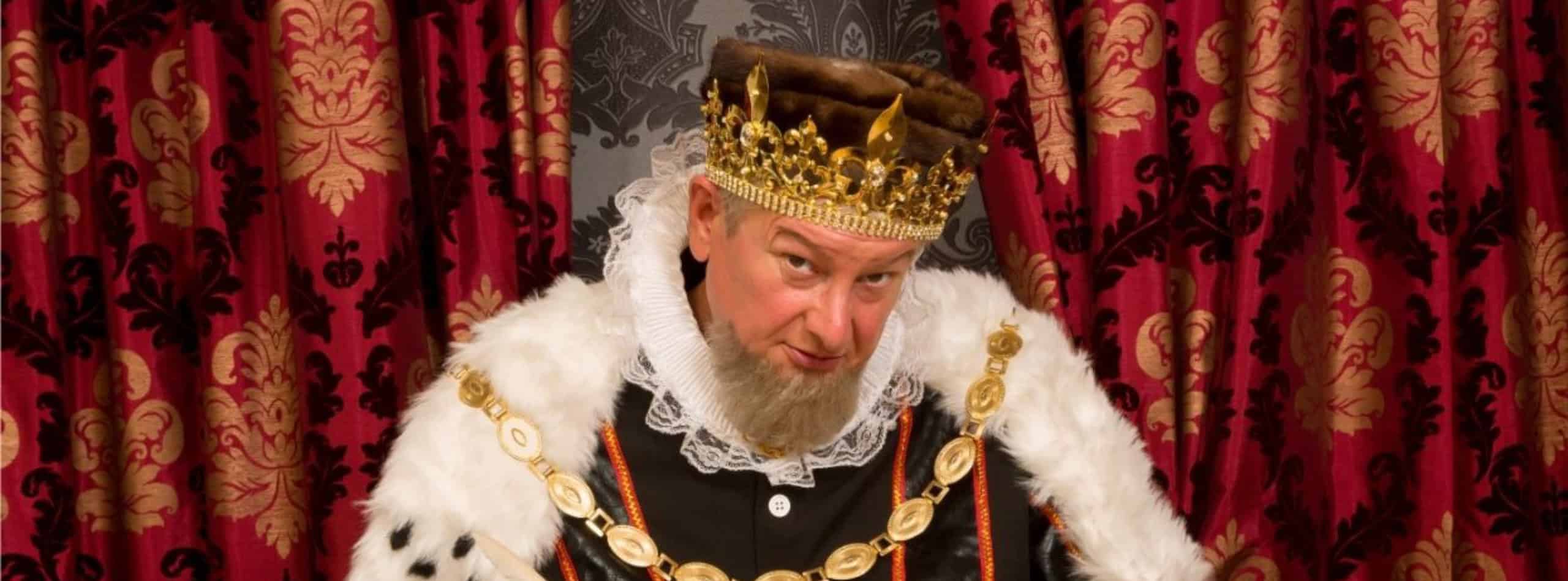 Ein als König gekleideter Mann, der die Anziehungskraft von Adelstitel kaufen - Deutsche Adelstitel verkörpert, sitzt mit Krone und pelzverbrämtem Gewand vor rot-goldenen Vorhängen und blickt ernst in die Kamera. | adelstitel-kaufen.com