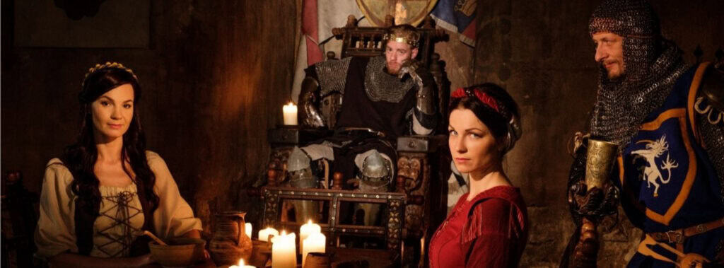 Vier Personen in mittelalterlichen Kostümen sitzen um einen mit Kerzen beleuchteten Tisch. Ein als König verkleideter Mann sitzt auf einem Thron, während zwei Frauen und ein Mann in einer Rüstung in der schummrigen Atmosphäre des Raumes über adelstitel kaufen - historische Informationen diskutieren. | adelstitel-kaufen.com