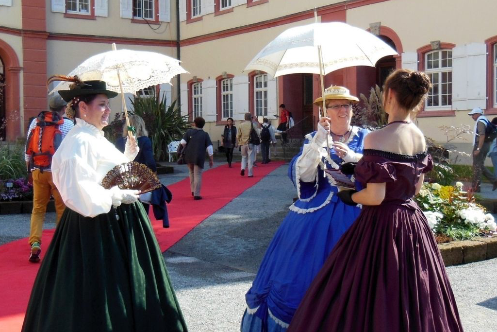 Drei Frauen in Kleidern und Hüten aus der viktorianischen Zeit stehen auf einem roten Teppich vor einem historischen Gebäude und unterhalten sich. Zwei Frauen halten weiße Sonnenschirme und eine einen Fächer, vielleicht diskutieren sie darüber, wie man inmitten der eleganten Menge und der Blumen Adelstitel kaufen kann. | adelstitel-kaufen.com
