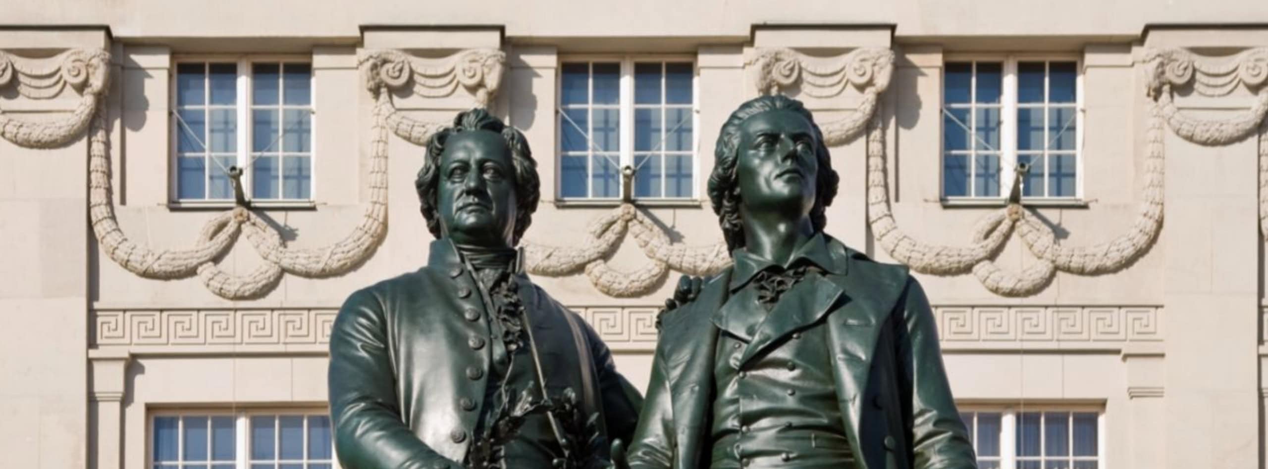 Zwei Bronzestatuen von Männern in historischen Gewändern stehen nebeneinander vor einem prunkvollen Gebäude, das die Zeit der Weimarer Verfassung widerspiegelt, mit dekorativen Girlanden und großen Fenstern, die die Szene aufwerten. | adelstitel-kaufen.com