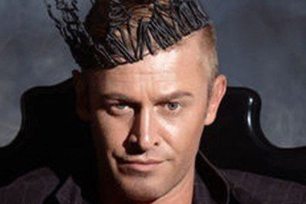 Eine Person mit kurzen blonden Haaren trägt eine schwarze Krone aus gedrehten Drähten, sitzt auf einem dunklen Stuhl und blickt mit einem leichten Lächeln direkt in die Kamera, als sei sie bereit, faszinierendes Wissenswertes zu vermitteln. | adelstitel-kaufen.com