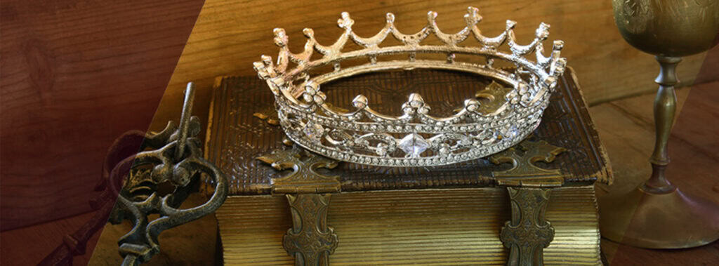 Eine silberne, juwelenbesetzte Krone ruht auf einem verschnörkelten, antiken Buch mit Metallklammern - Symbole des Ehrenkodex des Adels. Daneben liegen ein alter Schlüssel und ein goldener Kelch vor einem hölzernen Hintergrund. | adelstitel-kaufen.com