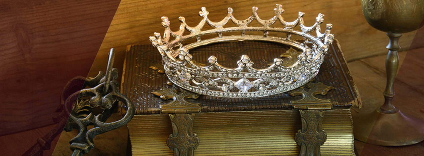 Eine silberne, juwelenbesetzte Krone ruht auf einem verschnörkelten, antiken Buch mit Metallklammern - Symbole des Ehrenkodex des Adels. Daneben liegen ein alter Schlüssel und ein goldener Kelch vor einem hölzernen Hintergrund. | adelstitel-kaufen.com