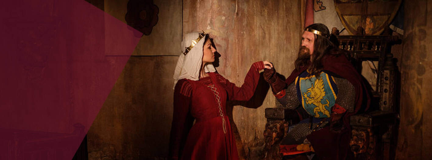 In einem schwach beleuchteten, rustikalen Raum mit Steinwänden hält eine Frau in einem mittelalterlichen roten Kleid und weißem Kopfschmuck einem sitzenden Mann die Hand, der eine Krone trägt - seine Rüstung zeigt einen Löwen, der die Adelstitel-Rangfolge widerspiegelt. | adelstitel-kaufen.com